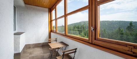A balcony with a wooden table and chairs, a view of a forested hillside, and a white textured wall.