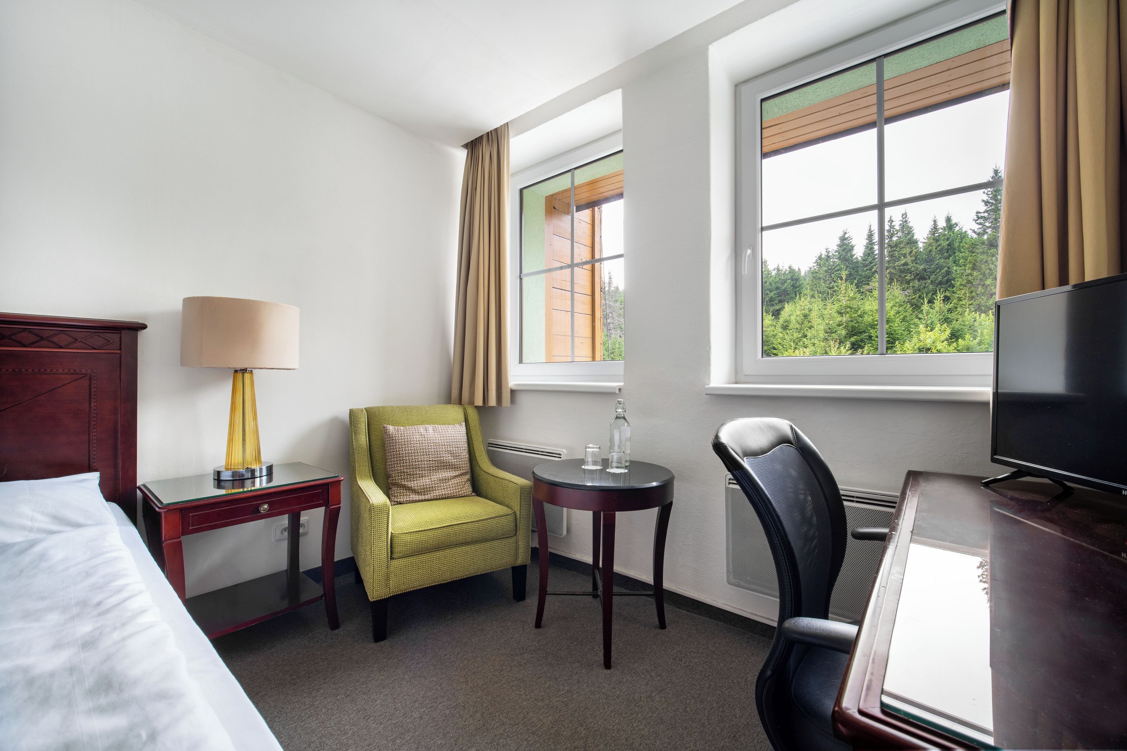A hotel room with a bed, a desk with a lamp, a green armchair, a small round table with two glasses, a television, and a window with curtains.