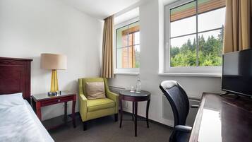 A hotel room with a bed, a desk with a lamp, a green armchair, a small round table with two glasses, a television, and a window with curtains.