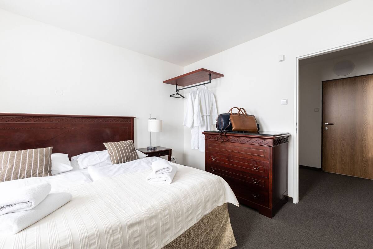 A neatly made bed with white linens, a wooden dresser with drawers, a hanging coat rack, and a door to the right.