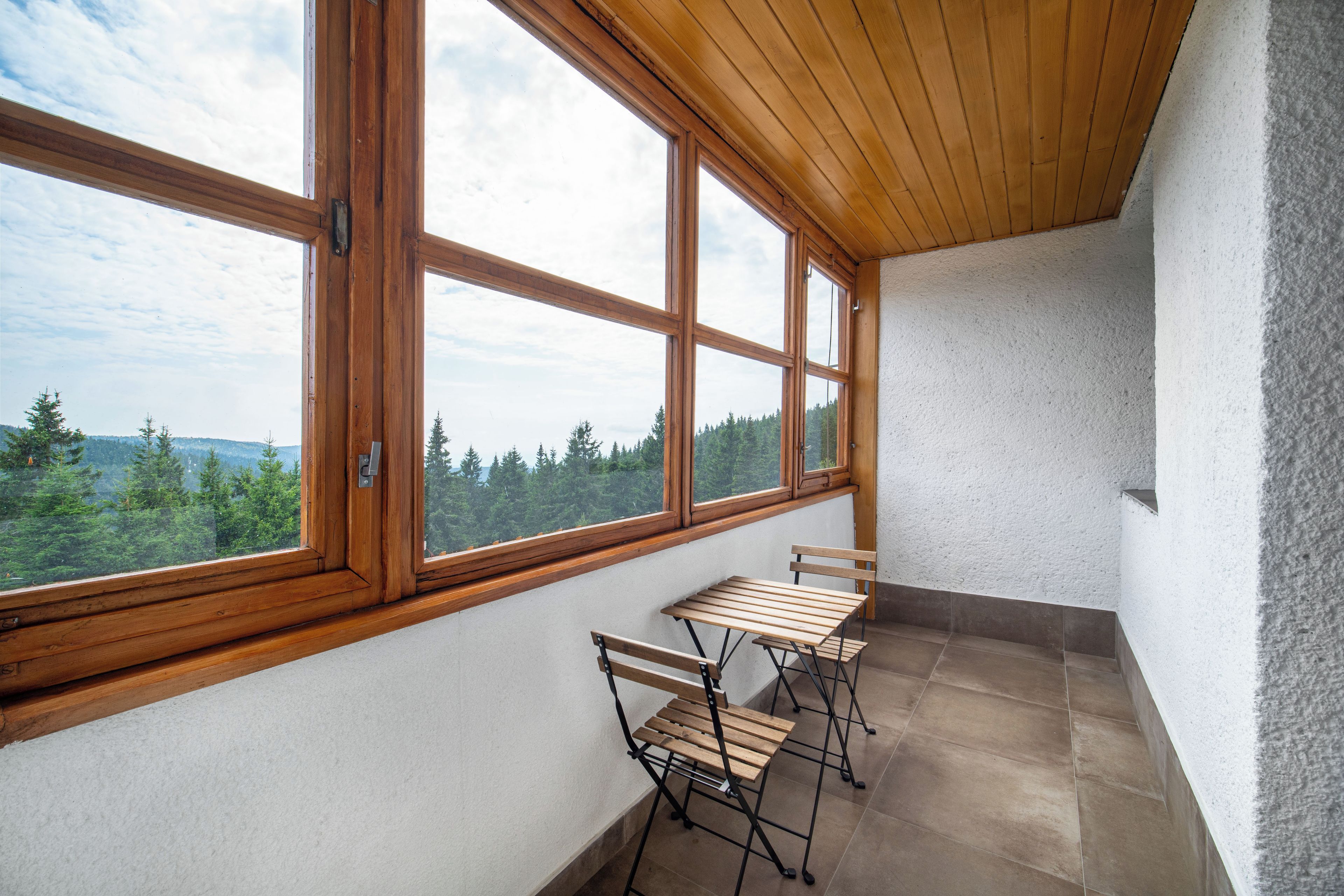 A balcony with a wooden table and chairs, large windows with a view of trees and mountains, and a textured white wall.