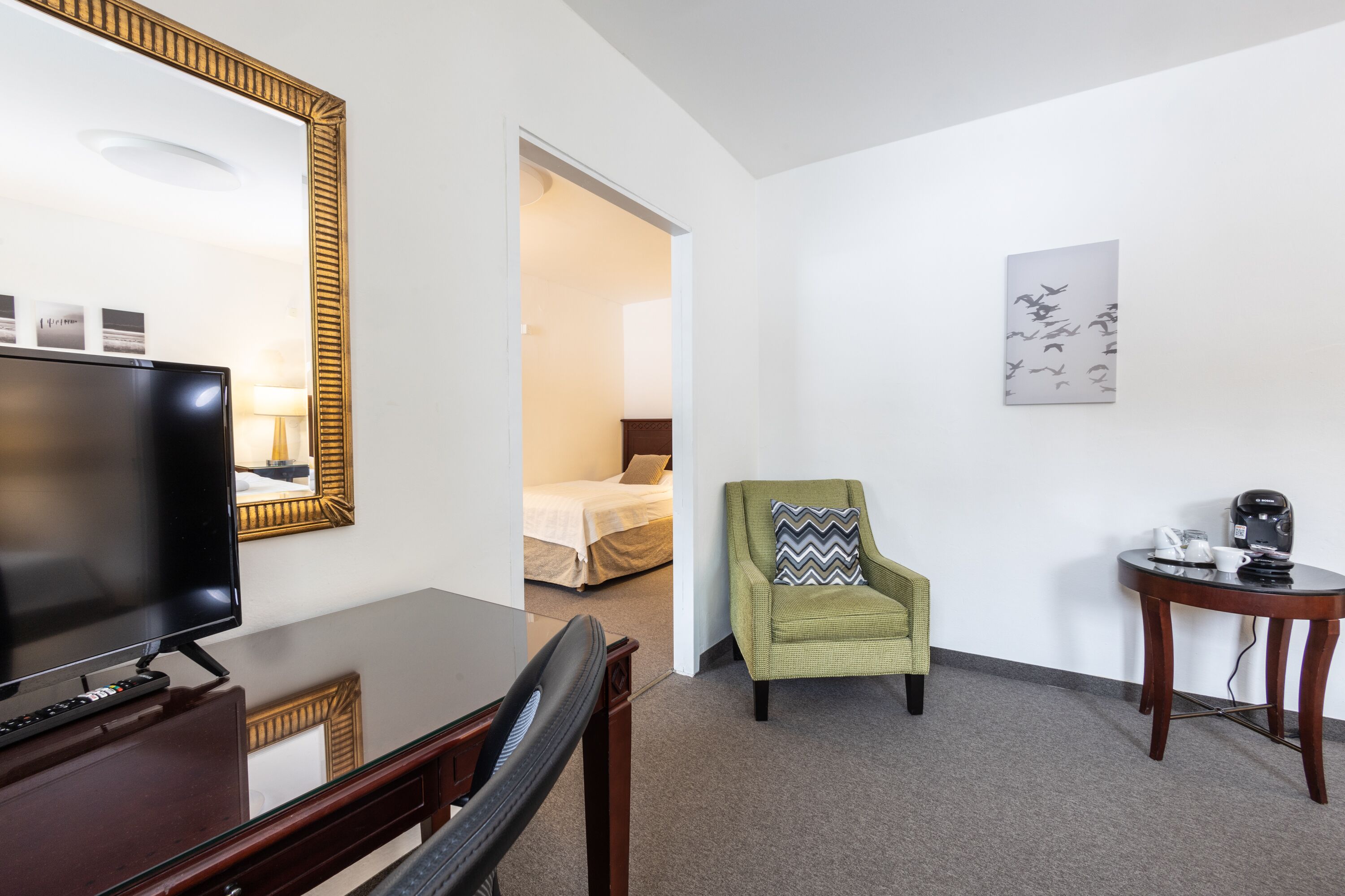 Une chambre d’hôtel équipée d’une télévision, d’un miroir, d’un fauteuil vert, d’une petite table avec une machine à café et d’un tableau au mur.