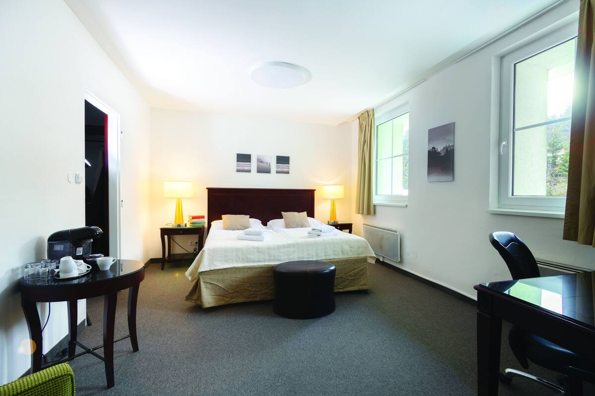 A hotel room with a bed, a desk, a chair, a coffee machine, and a lamp.