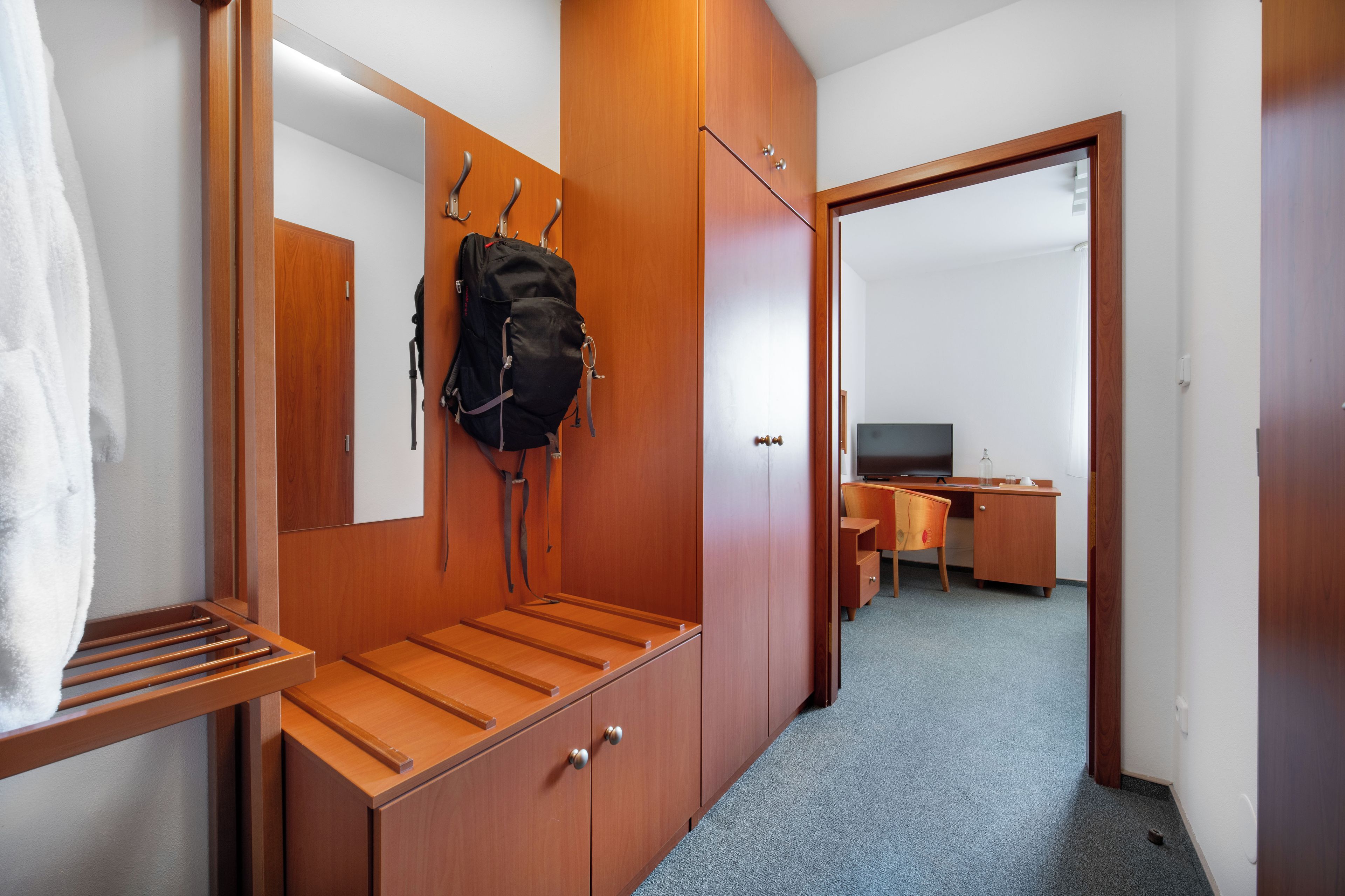 A hotel room with a wooden wardrobe, a mirror, a desk with a chair, and a television.