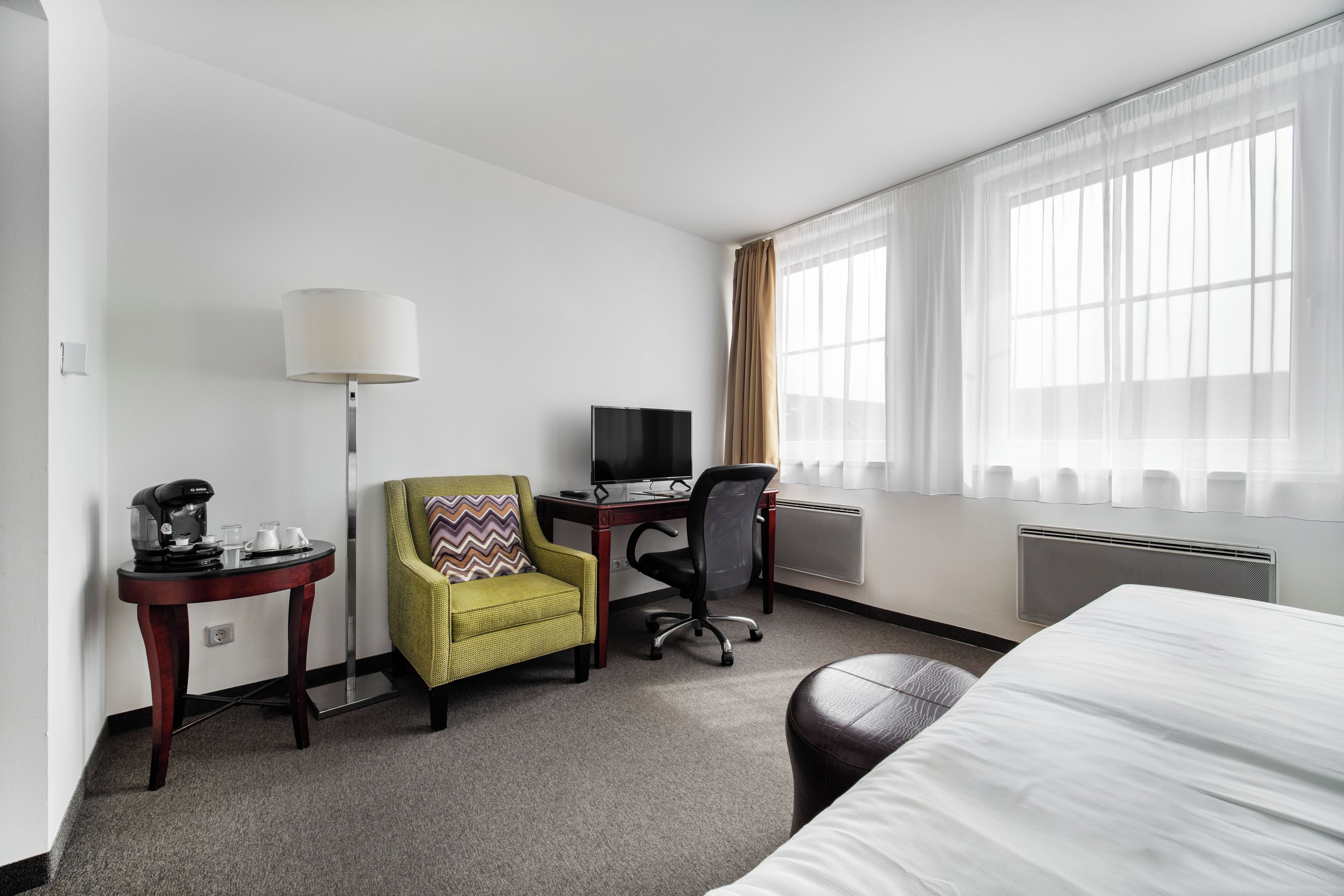 Quarto de hotel com cama, escrivaninha, computador, cadeira, luminária e cafeteira.