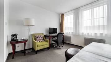 Quarto de hotel com cama, escrivaninha, computador, cadeira, luminária e cafeteira.