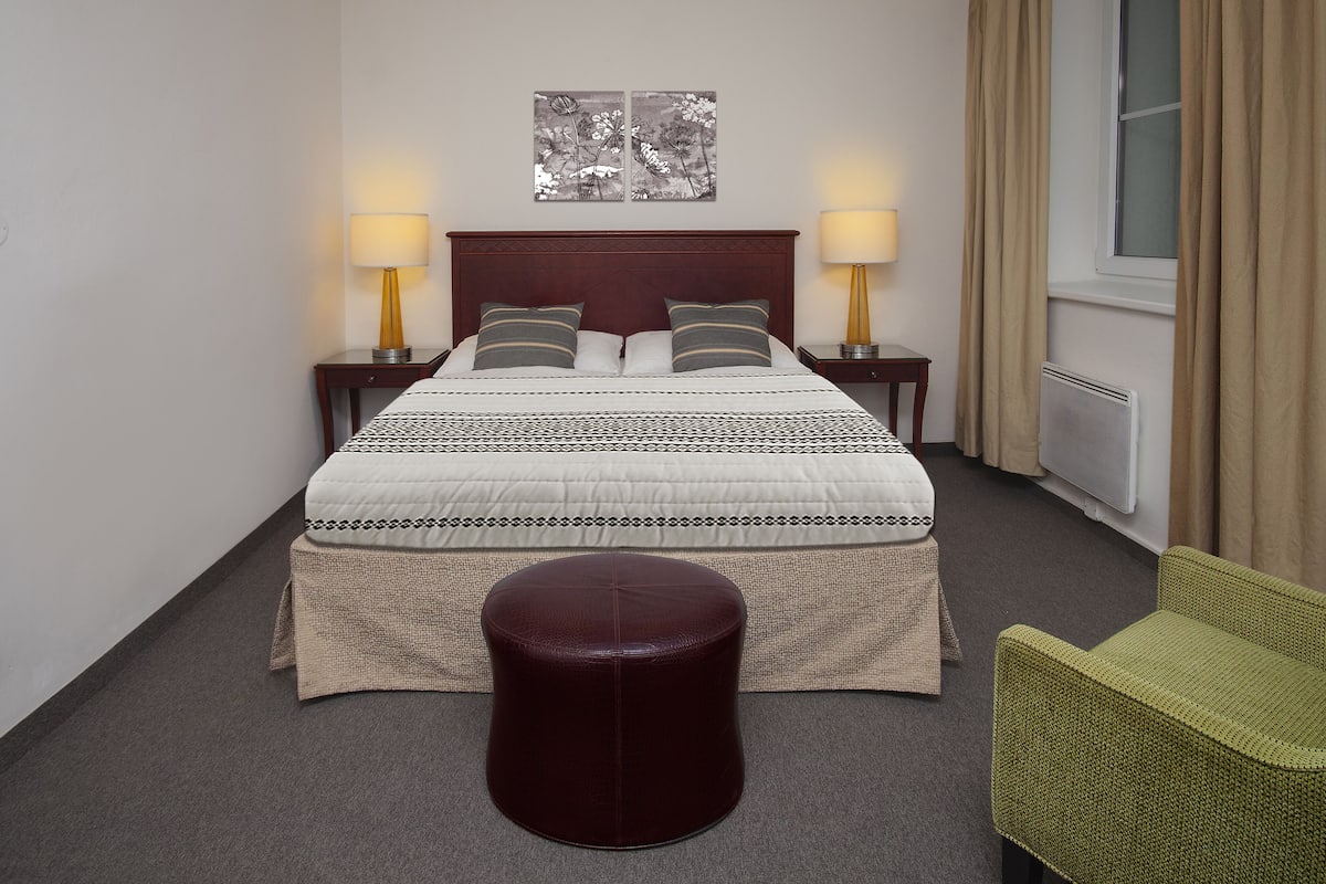 A hotel room with a bed, two bedside lamps, a window with curtains, a radiator, a green chair, and a round ottoman.