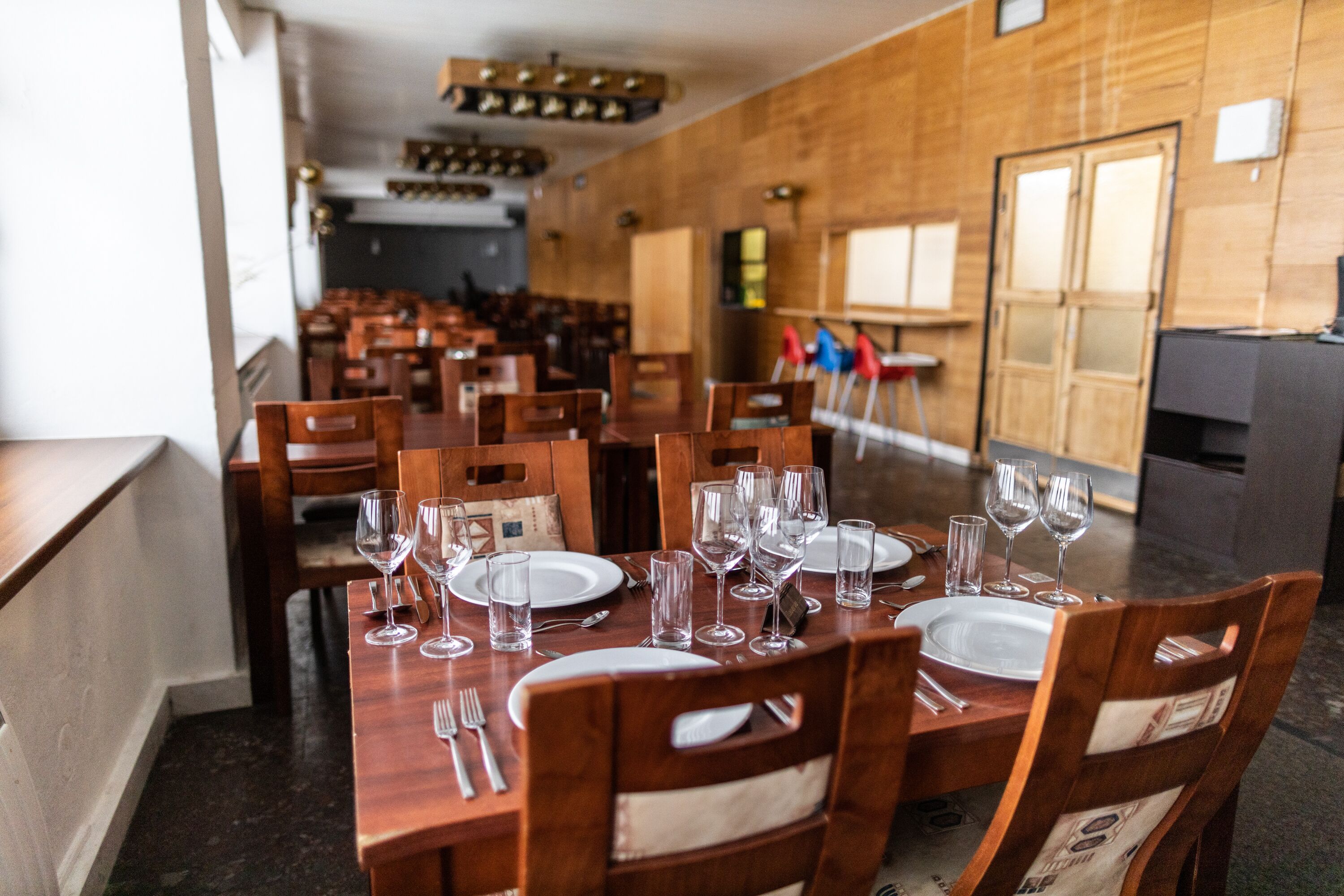 Un comedor con mesas y sillas de madera, cada juego con platos, cubiertos y copas de vino.