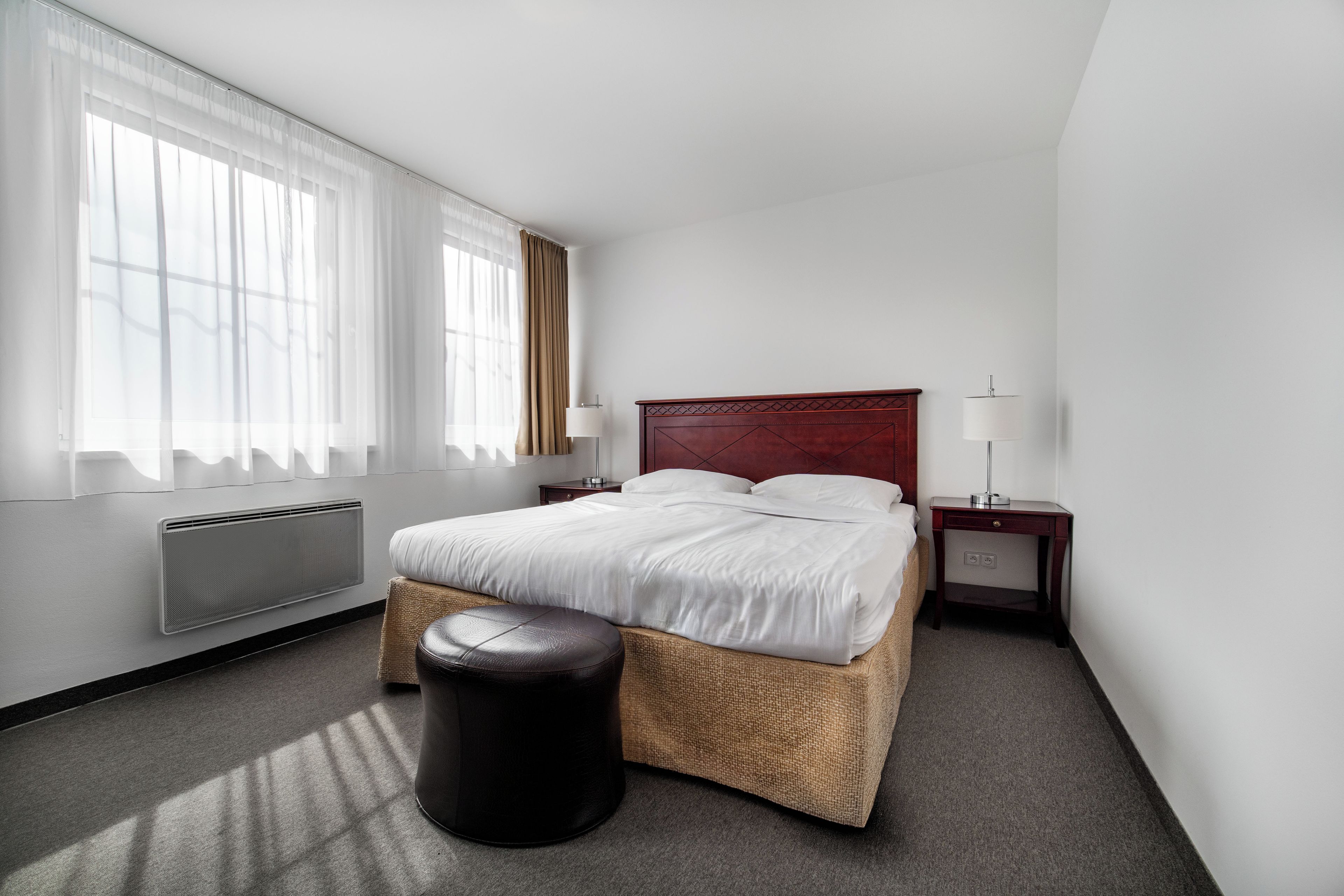 Een hotelkamer met een bed, een nachtkastje met een lamp, een radiator en een raam met gordijnen.