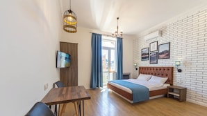 Una habitación de hotel moderna con suelo de madera, cama con cubrecama azul, mesa de comedor con sillas, televisor montado en la pared y candelabro.