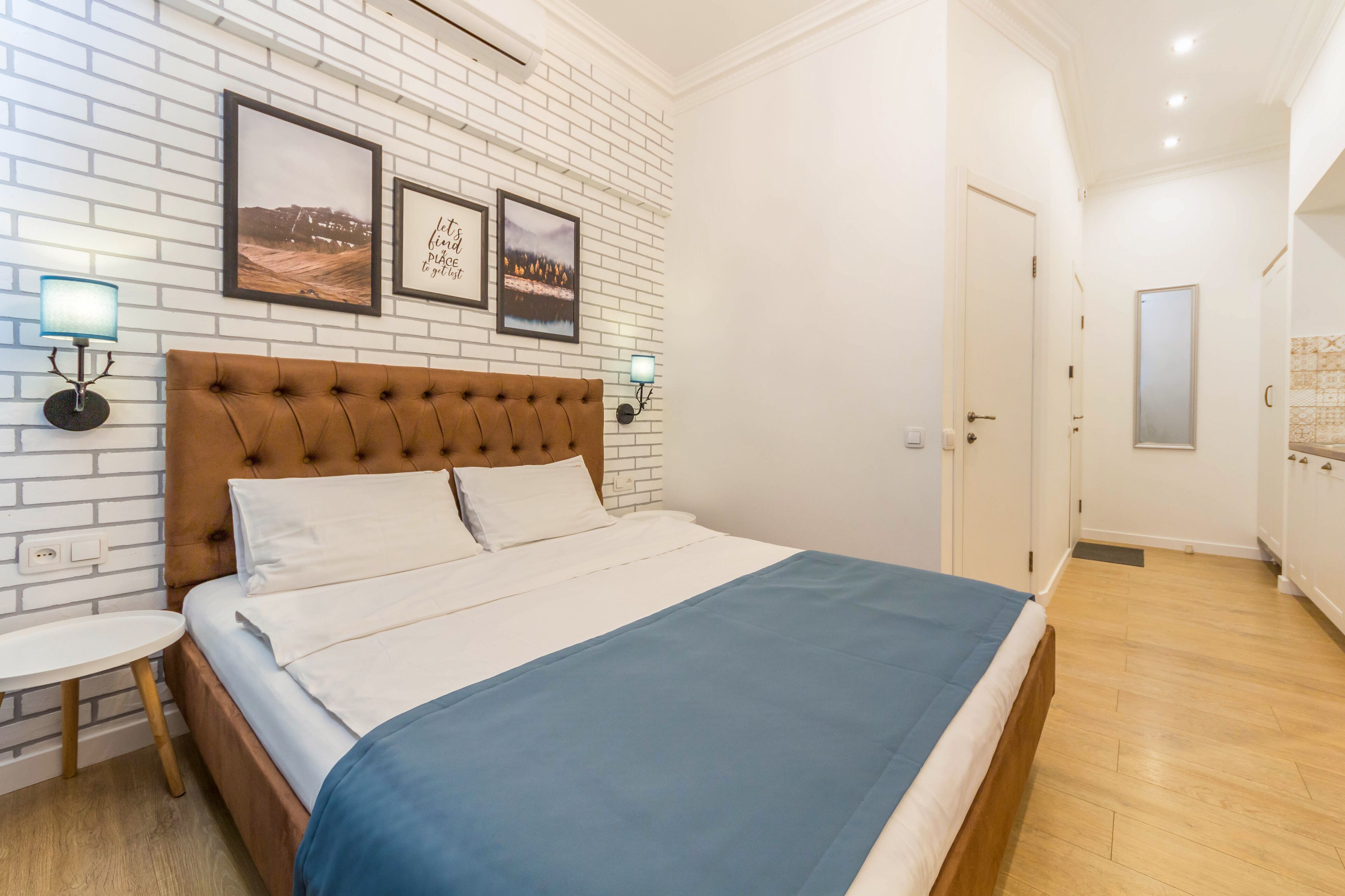 A bedroom with a large bed, white brick wall, two framed pictures, bedside lamps, and a small round table.