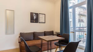 Un moderno salón con sofá, mesa de comedor y sillas. Se observa una araña, un amplio ventanal con cortinas y un cuadro enmarcado en la pared.