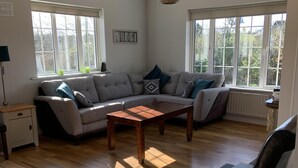 Sala de estar con un sofá seccional gris, una mesa de centro de madera y un sillón de cuero negro. Hay dos ventanas con marcos blancos y vistas a árboles desde el exterior.