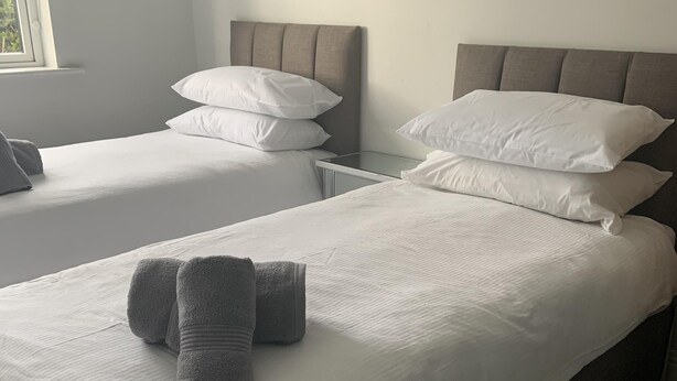 Habitación de hotel con dos camas, cada una con sábanas y almohadas blancas, un cabecero gris y una mesita de noche pequeña.