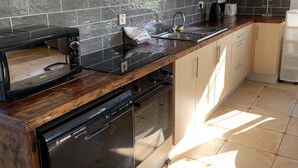 Una cocina con lavavajillas, estufa y microondas. Hay un fregadero con grifo y una cafetera. La cocina tiene paredes de azulejos y armarios de madera.