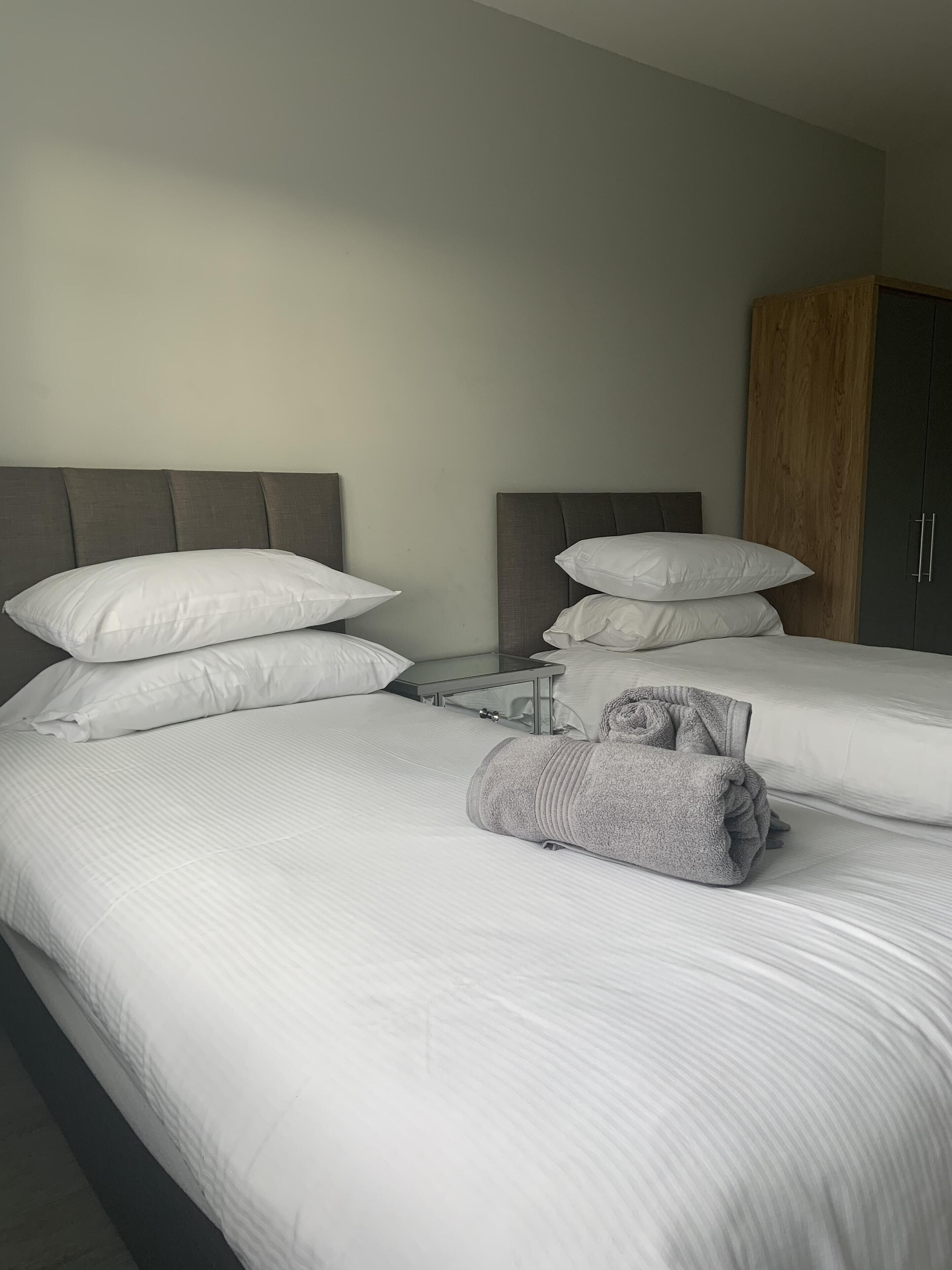 Habitación de hotel con dos camas, cada una con ropa blanca y almohadas. Se observa un armario de madera y una mesita con una lámpara.