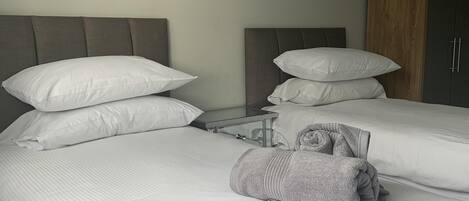 Habitación de hotel con dos camas, cada una con ropa blanca y almohadas. Se observa un armario de madera y una mesita con una lámpara.