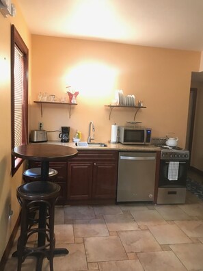Cozinha compacta com torradeira, micro-ondas, fogão e lava-louças. Há dois bancos e uma mesa pequena.