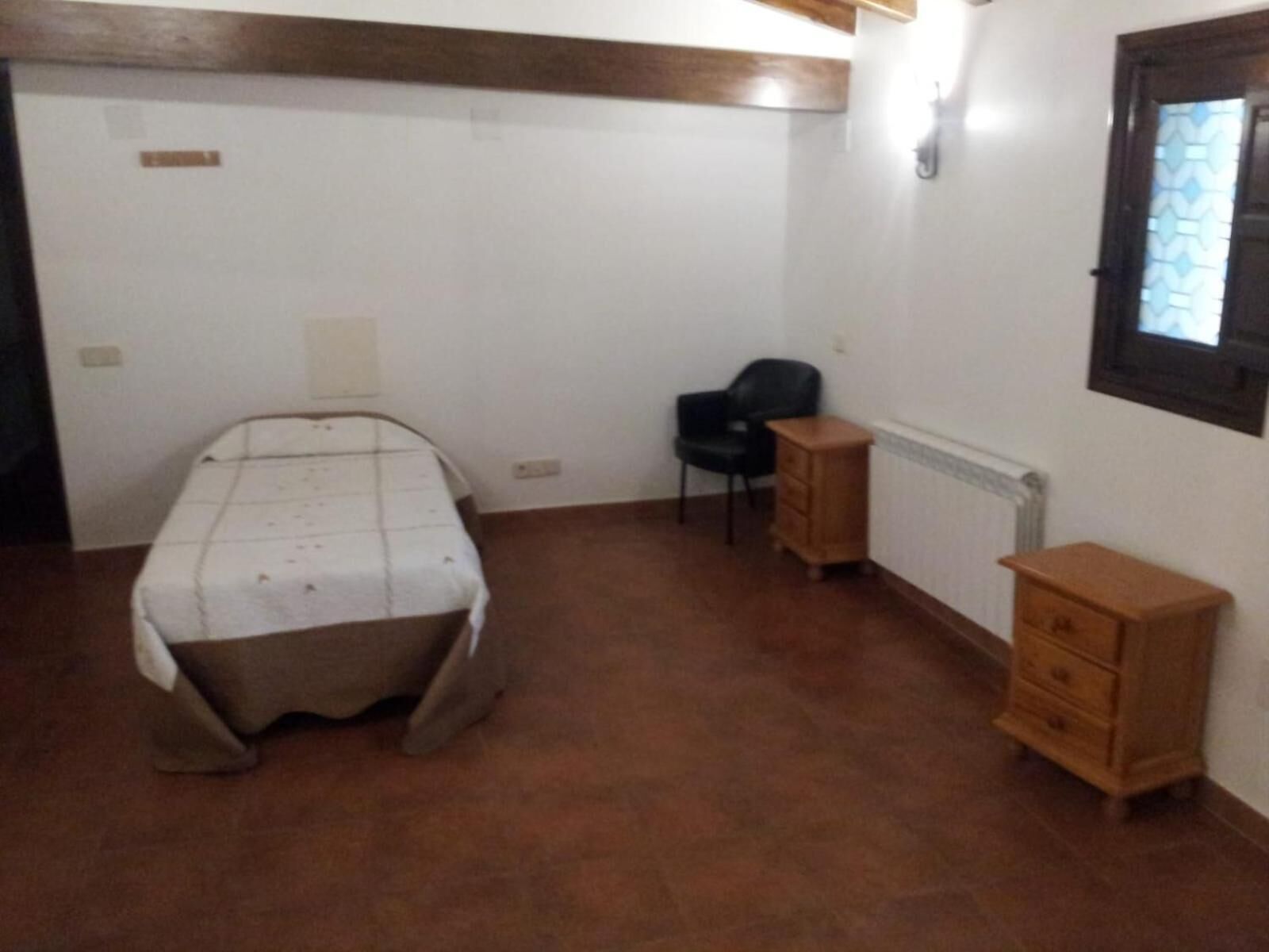 Una habitación pequeña y poco amueblada con una cama, una silla, un aparador de madera y un radiador.
