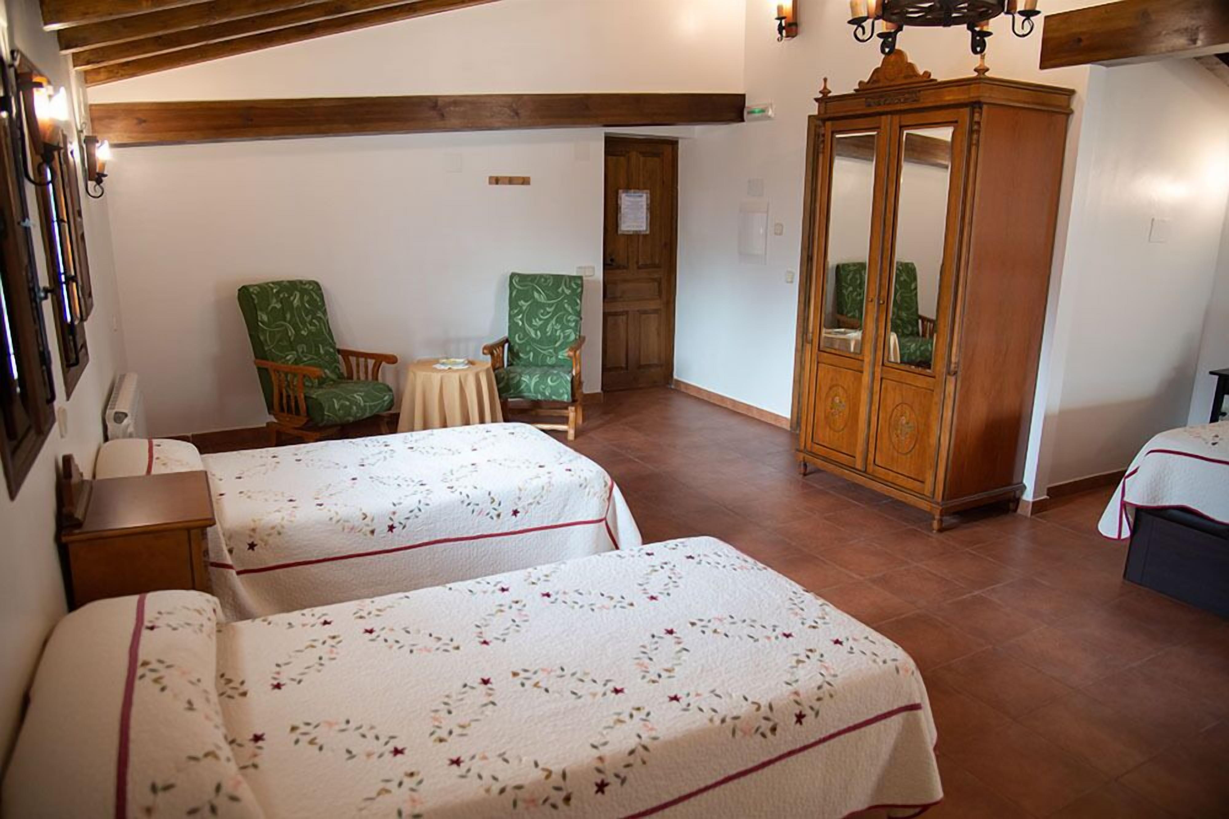 A room with two beds, a wooden wardrobe, and two green armchairs.