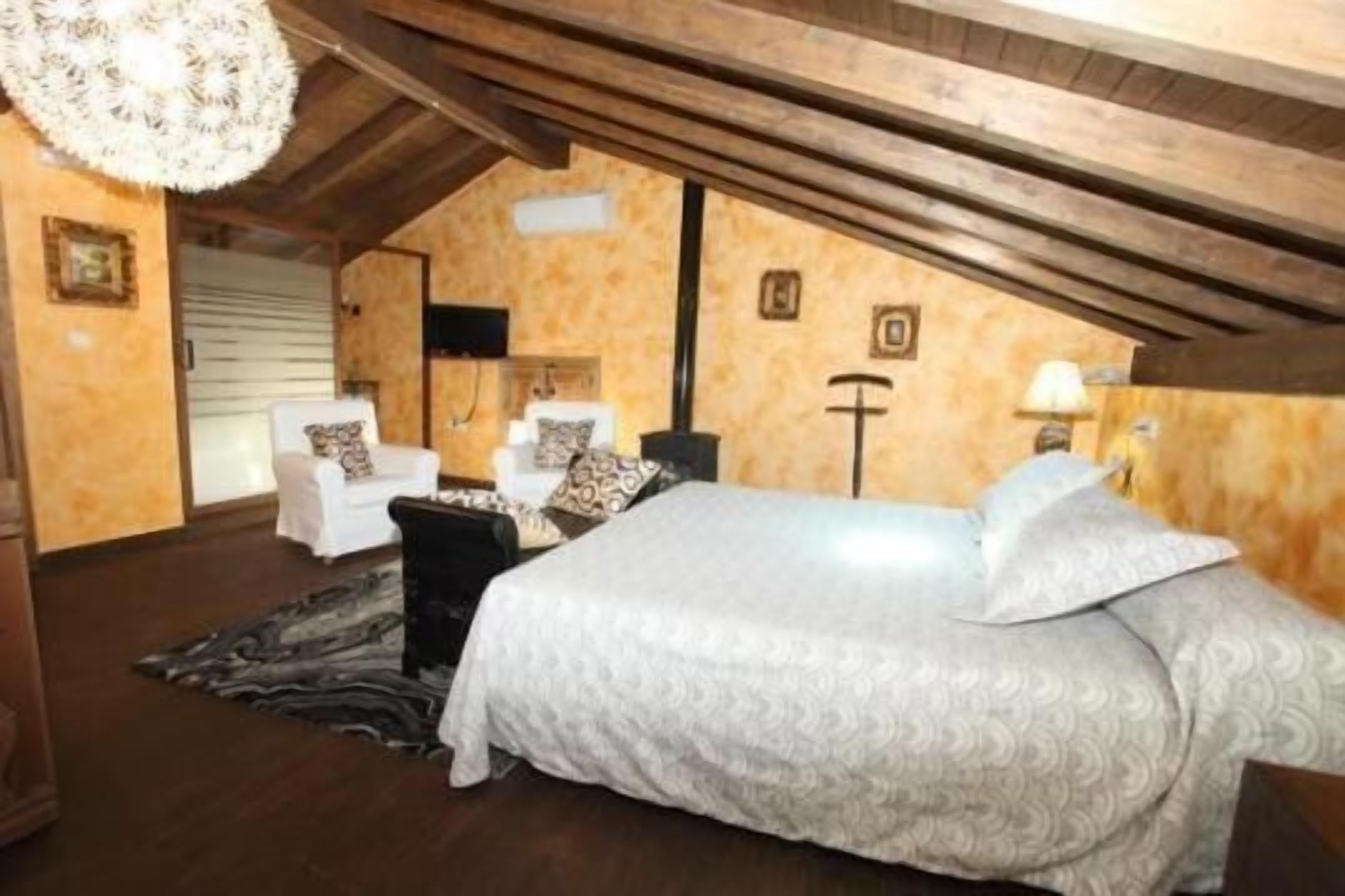 Une chambre mansardée chaleureuse, avec un plafond en pente, un lit recouvert d’une couvre-lit à motifs, un fauteuil blanc, une table de chevet noire et un lampadaire.