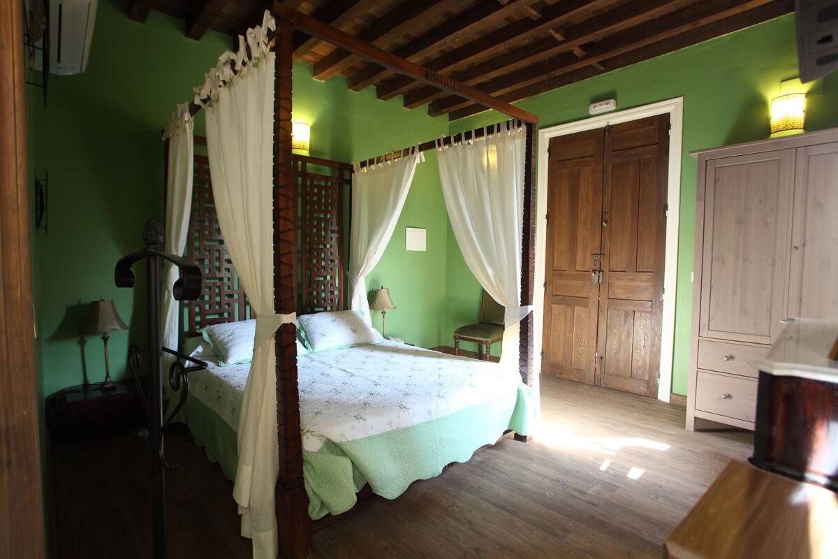 Una cama con dosel con cortinas blancas, un armario de madera y una mesita de noche con lámpara.