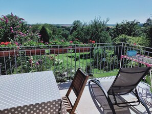 Une terrasse avec une table et des chaises, entourée d’une clôture en métal, donnant sur un jardin où poussent différentes plantes et fleurs.