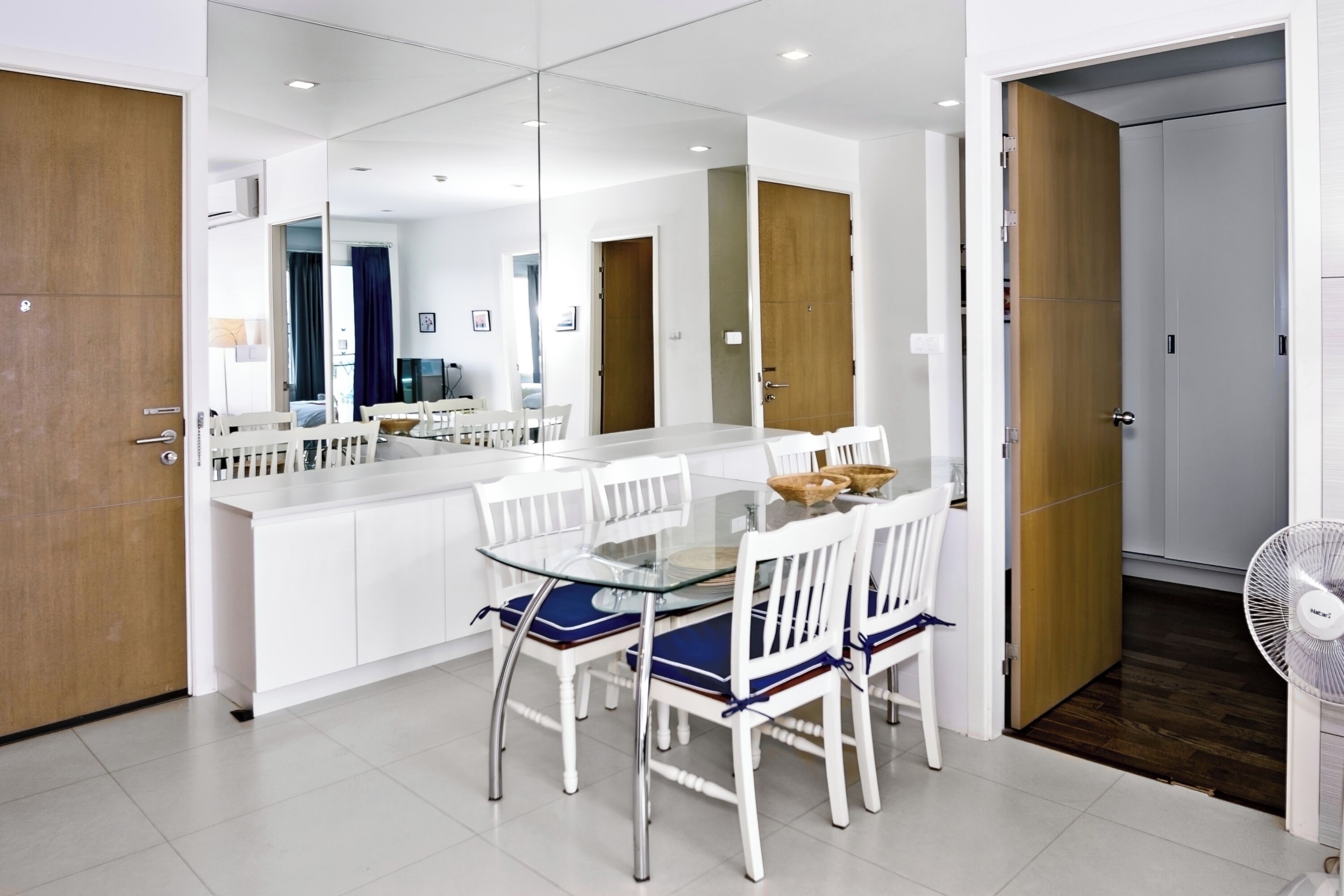 Un espace repas moderne avec une table en verre et des chaises blanches, une porte en bois et un ventilateur blanc.