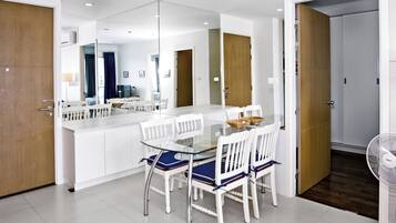 Un espace repas moderne avec une table en verre et des chaises blanches, une porte en bois et un ventilateur blanc.