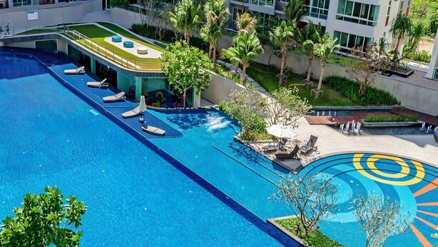 A large swimming pool with multiple lounge chairs and a sunbathing area, surrounded by palm trees and greenery. - SEACRAZE D709 - 2 Bedroom - Near Beach (Hua Hin)