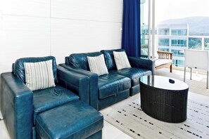 Ein modernes Wohnzimmer mit einem blauen Ledersofa, einem runden schwarzen Couchtisch und Blick auf ein Gebäude durch große Fenster.