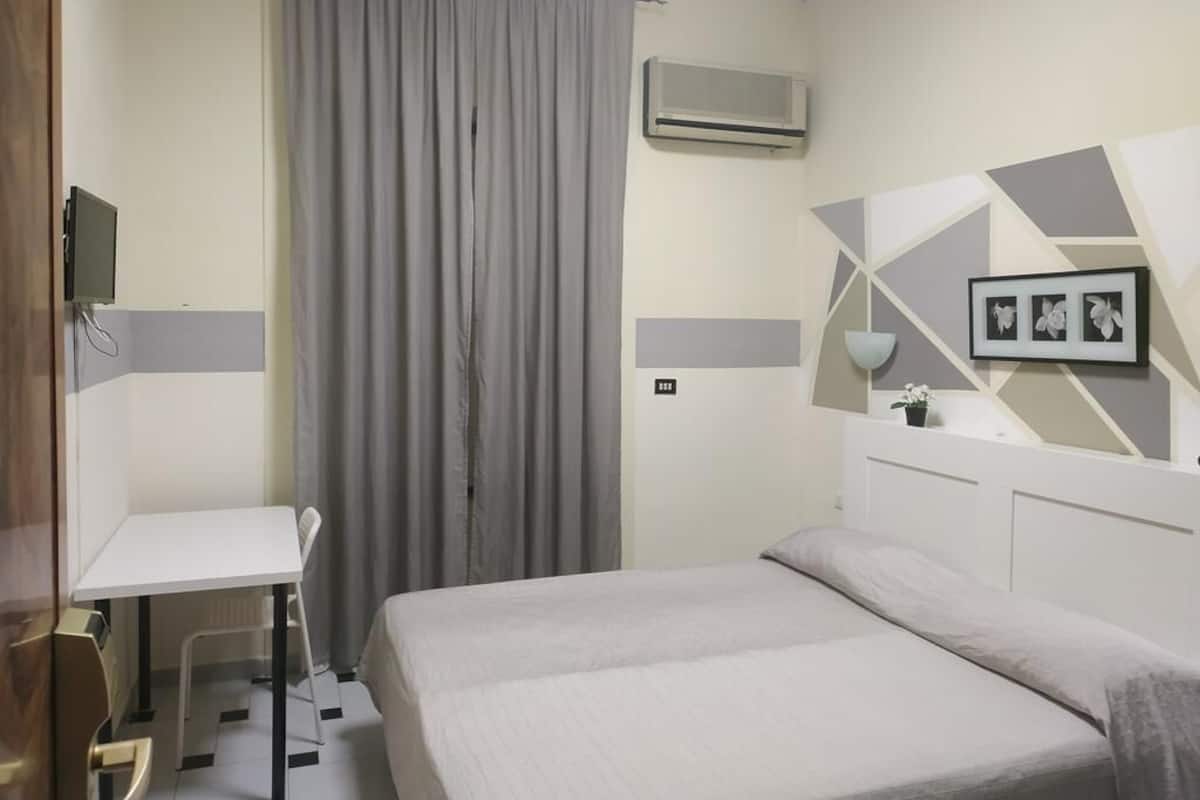 A hotel room with a bed, a desk, and a chair. There is a curtain, an air conditioning unit, and a wall-mounted shelf with a plant.