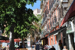 Une rue étroite bordée d’immeubles à plusieurs étages, dont certains sont dotés de balcons et d’espaces aménagés pour s’asseoir à l’extérieur.