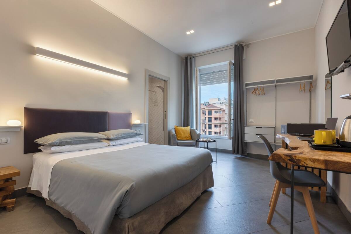 Ein modernes Hotelzimmer mit einem großen Bett, einem Schreibtisch mit einem Laptop, einem Stuhl und einem Fenster mit Blick auf Gebäude.