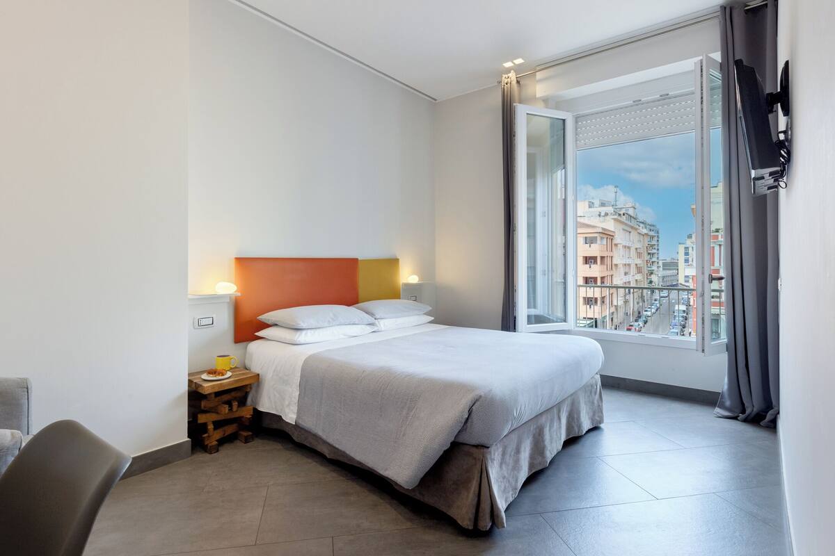 Ein modernes Hotelzimmer mit einem großen Bett, einem Nachttisch mit Lampe, einem Sessel und Blick auf die Stadt durch ein großes Fenster.