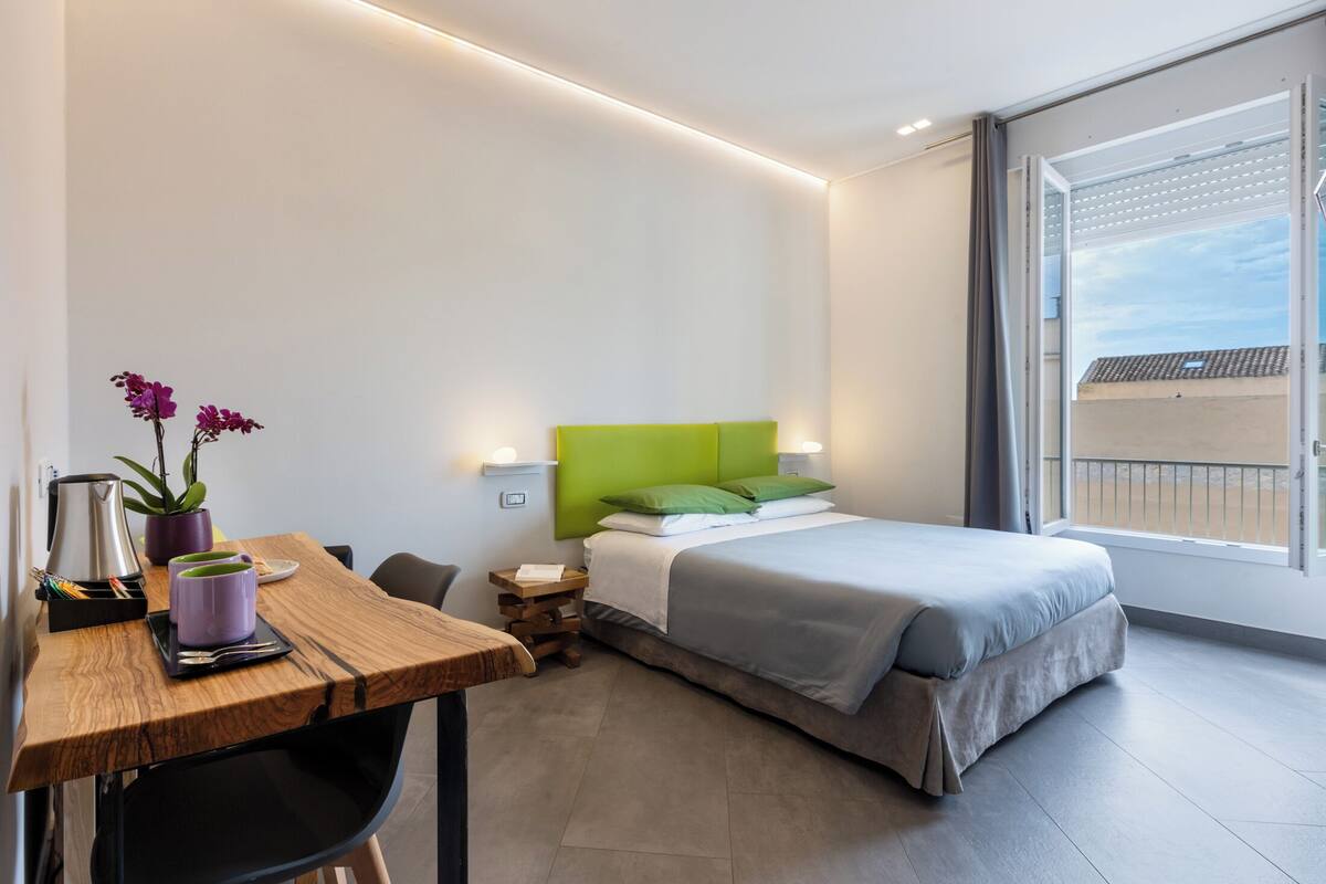 Ein modernes Hotelzimmer mit Bett, Schreibtisch mit Stuhl, einem kleinen Tisch, einer Zimmerpflanze und Blick nach außen.