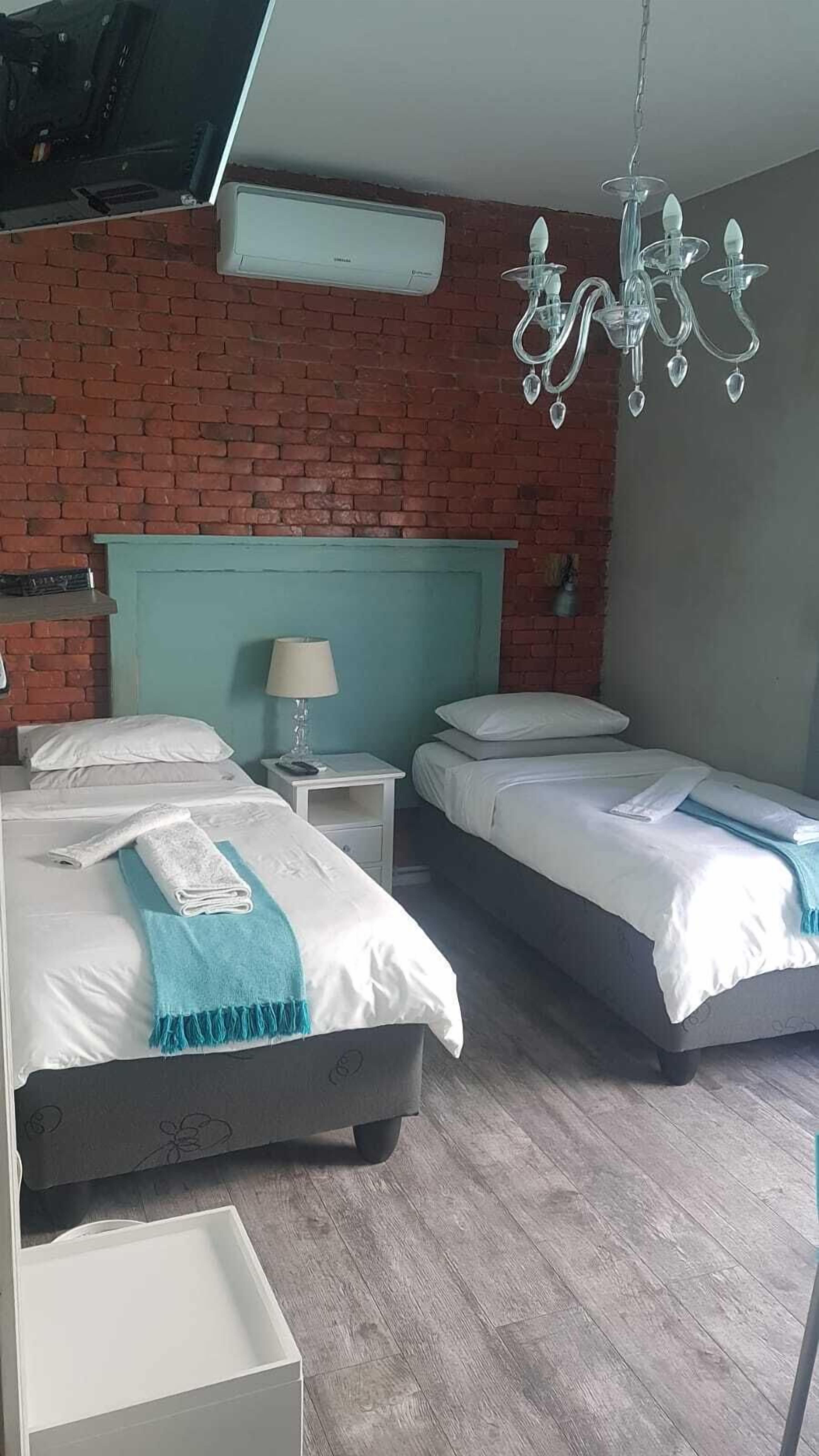 Quarto de hotel com duas camas, parede de tijolos, lustre e mesa de cabeceira com abajur.
