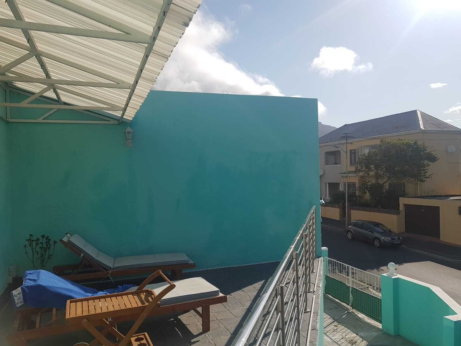 Un balcon avec un mur bleu, un fauteuil de jardin en bois, une chaise longue et une rambarde en métal.