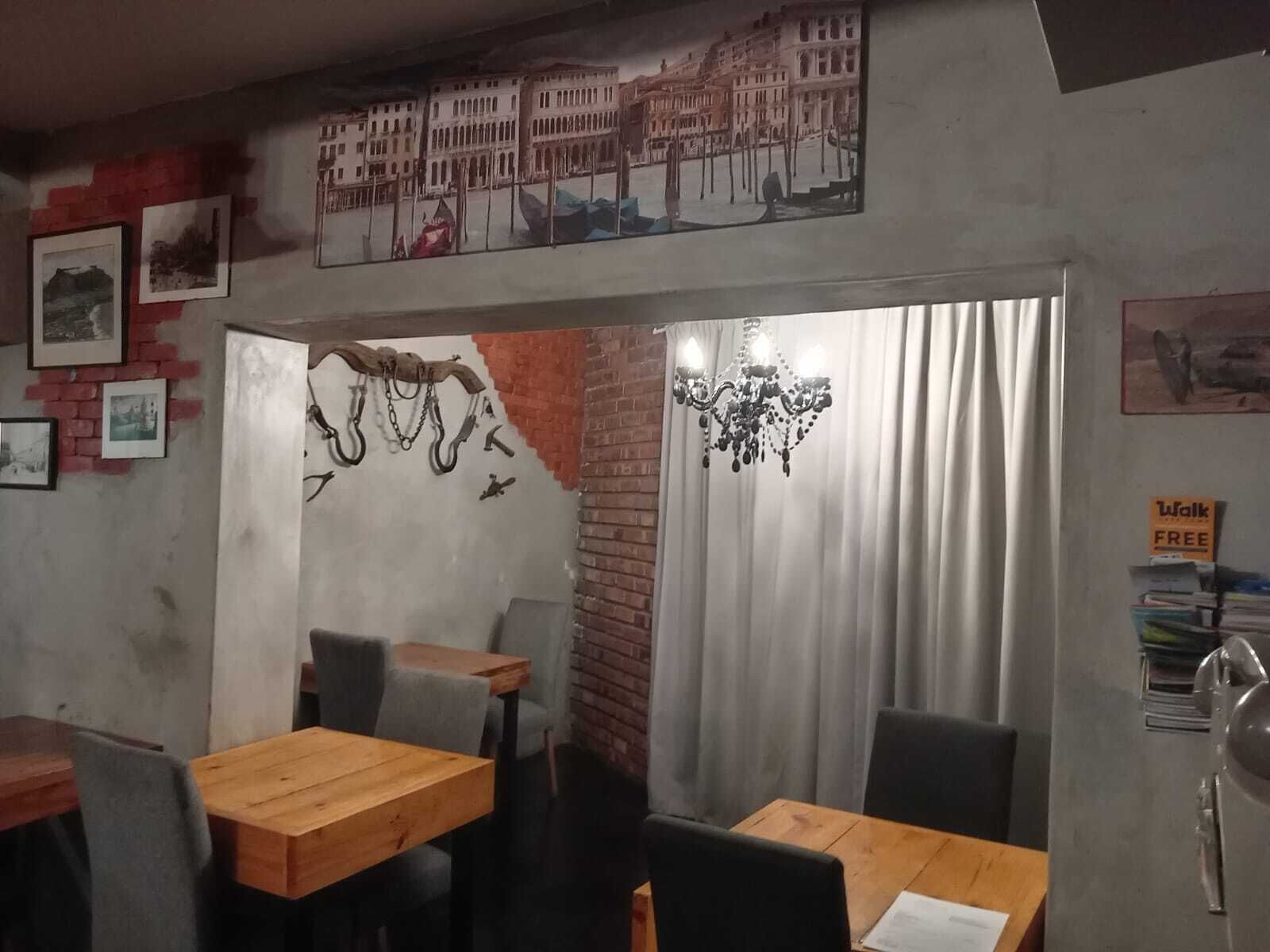 Een restaurant met bakstenen muren, houten tafels en stoelen. Er hangen ingelijste foto’s aan de muren en er is een kroonluchter.