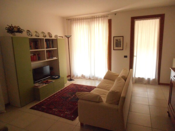 A living room with a beige sofa, a green entertainment unit with a TV, a red rug, and a large window with curtains.