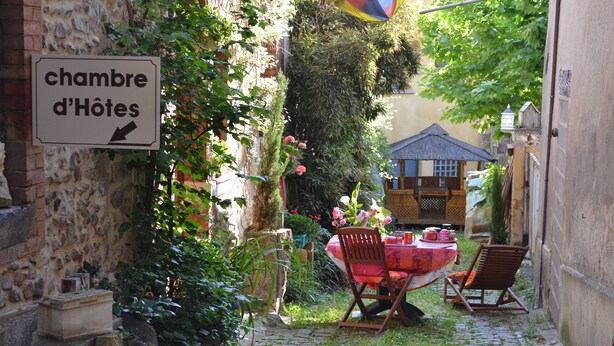 Un camino empedrado que lleva a un jardín con una mesa y sillas, un cartel que dice 'chambre d'Hôtes', y un edificio al fondo.