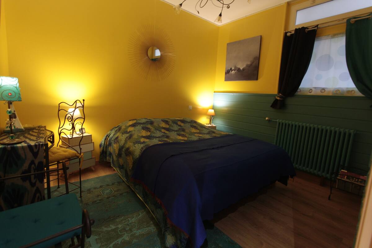 Una habitación con cama, escritorio, silla, lámpara, radiador y ventana con cortinas.