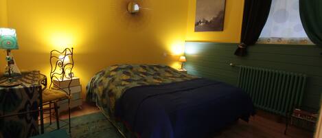 Una habitación con cama, escritorio, silla, lámpara, radiador y ventana con cortinas.