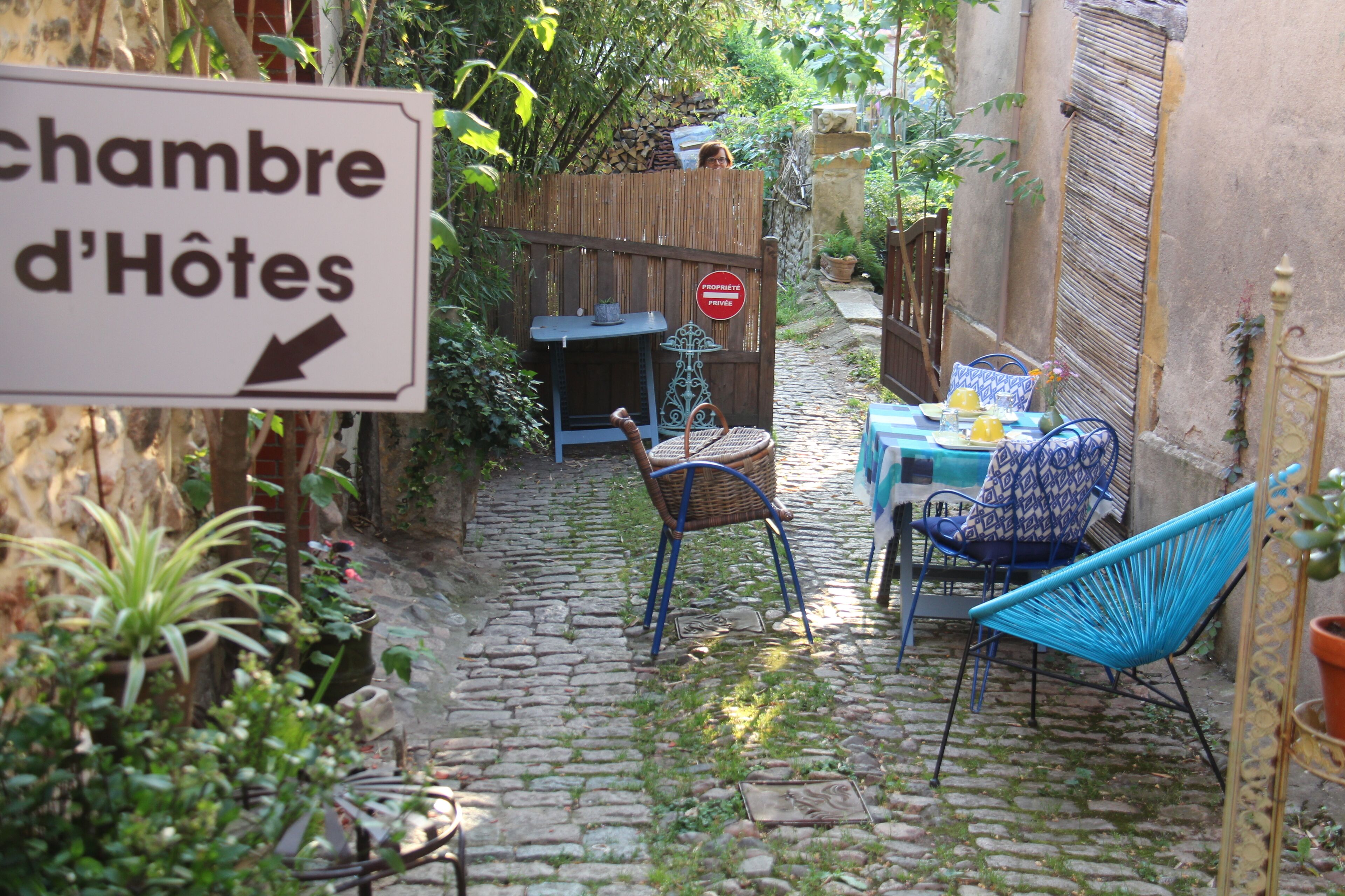 A narrow cobblestone alley with a sign for 'chambre d'Hôtes' pointing to the right. There are two wicker chairs and a table with a blue chair.