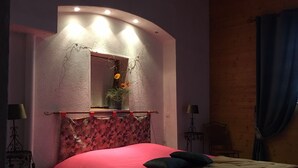 Un dormitorio con cabecera curvada, cama con colcha roja y marrón, un espejo con un florero y dos mesitas de noche con lámparas.