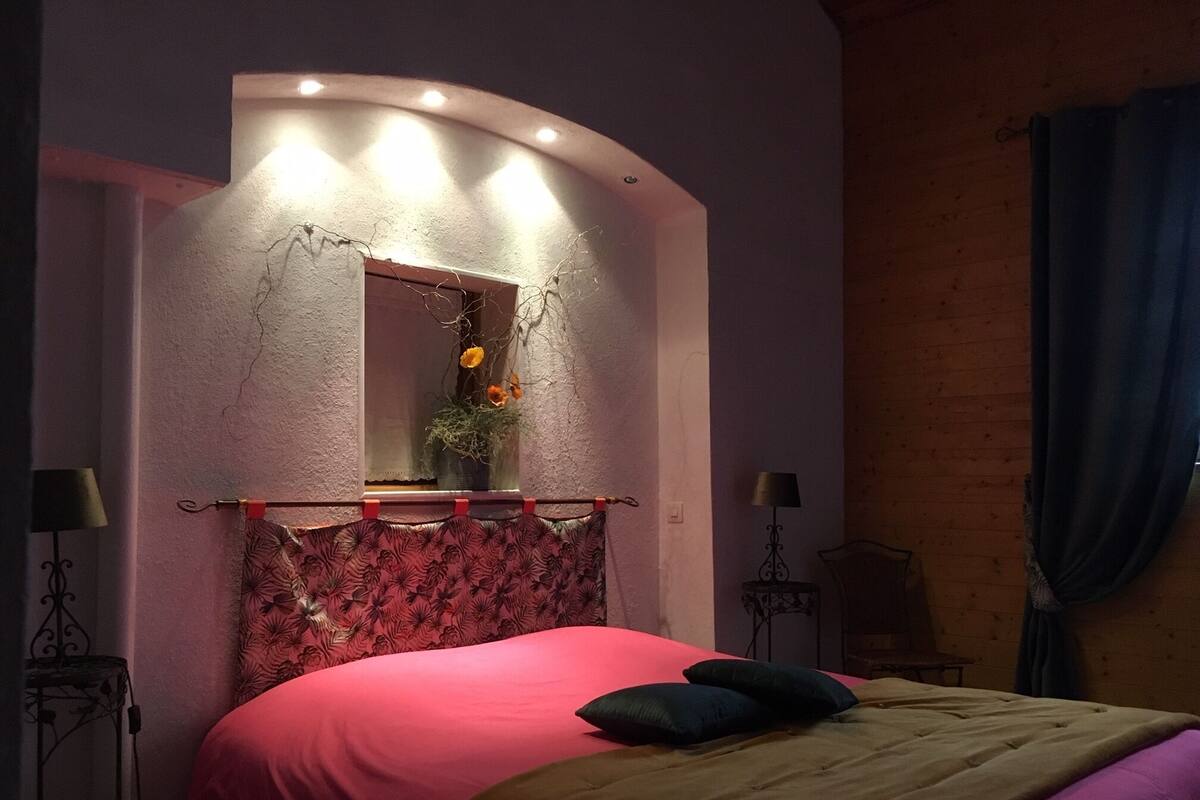 Un dormitorio con cabecera curvada, cama con colcha roja y marrón, un espejo con un florero y dos mesitas de noche con lámparas.