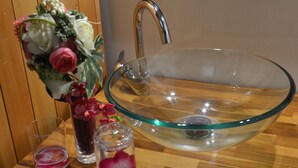 Un lavamanos de cristal transparente con un grifo moderno, un florero con flores rosas y dos vasos pequeños sobre una superficie de madera.