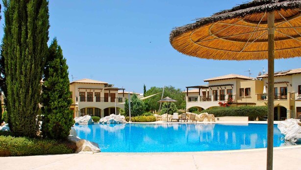Una piscina con sillas de descanso y sombrillas, rodeada de edificios con balcones y un cielo despejado.