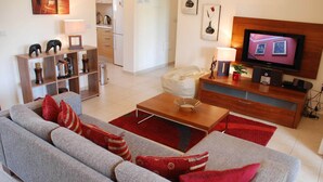 Una sala de estar moderna con un sofá gris, cojines rojos, una mesa de centro de madera y un televisor de pantalla plana sobre un mueble de madera.