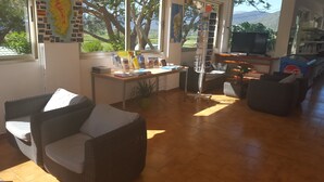 A cozy living room with wicker furniture, a desk with books and a computer, and a large window with a view of greenery and mountains. - Camping U Sommalu (Casaglione)